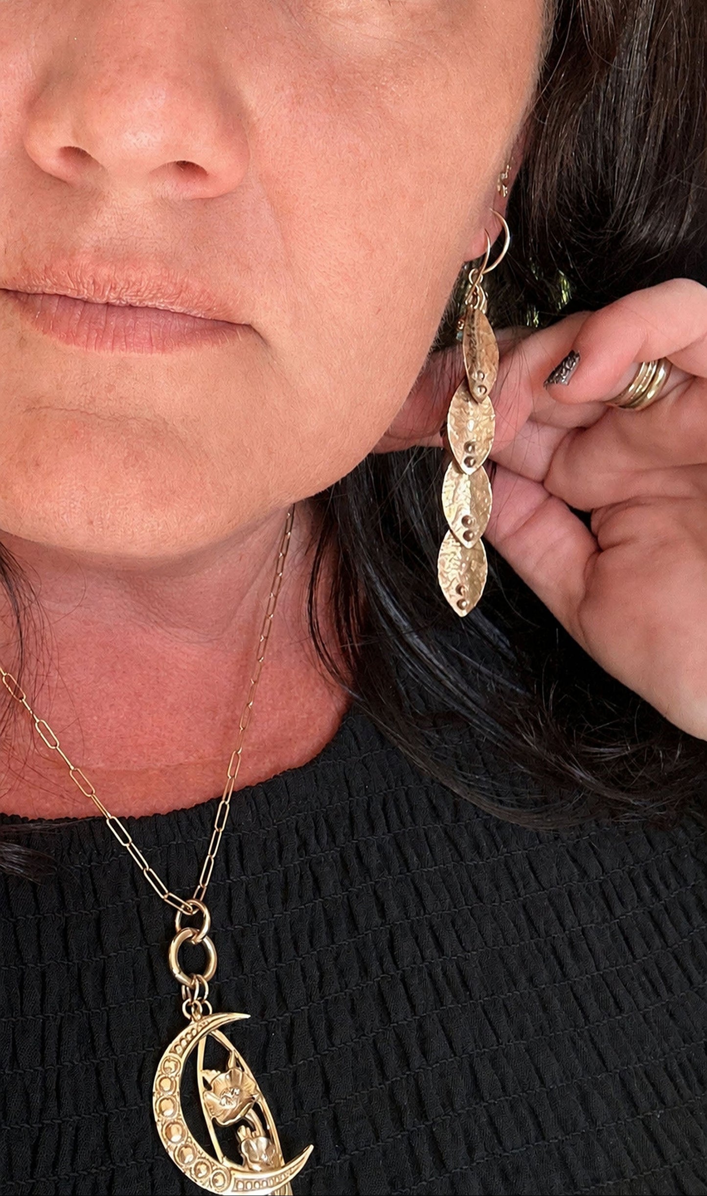 
                  
                    A close-up lifestyle shot of a woman wearing a gold crescent moon pendant necklace and matching gold dangle earrings, adjusting one of the earrings with her hand.
                  
                